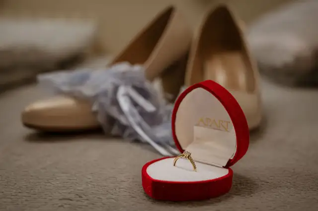 Złoty pierścionek zaręczynowy w czerwonym pudełku na tle butów ślubnych.