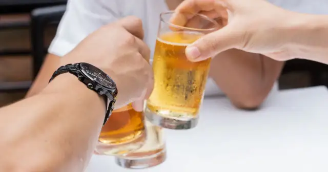 Dłonie unoszące szklanki z piwem do toastu, jedna z nich ma zegarek.