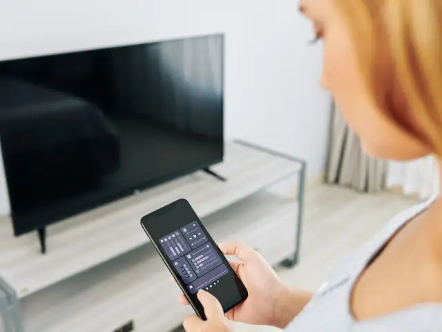 Kobieta trzyma smartfon z aplikacją do sterowania TV, obok stoi włączony telewizor.