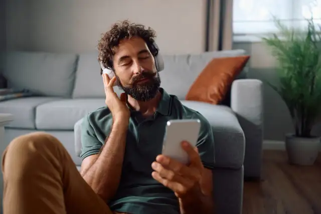 Mężczyzna w słuchawkach z telefonem w ręku, zrelaksowany na podłodze obok kanapy.