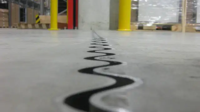 Szczegółowe wykonanie dylatacji w wylewce betonowej, widoczne na tle magazynowym z żółtymi słupami.