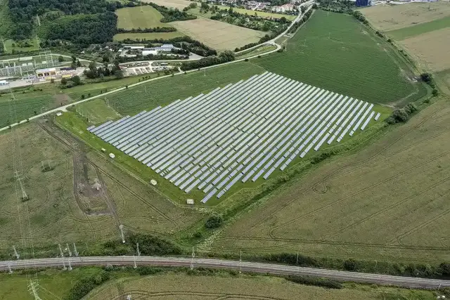 Widok z lotu ptaka na dużą farmę paneli słonecznych otoczoną polami i linią kolejową.