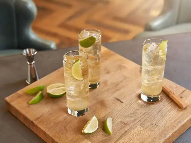 Trzy orzeźwiające whisky ze spritem, udekorowane plasterkami limonki, stoją na drewnianej desce. Obok leżą świeże kawałki limonki.