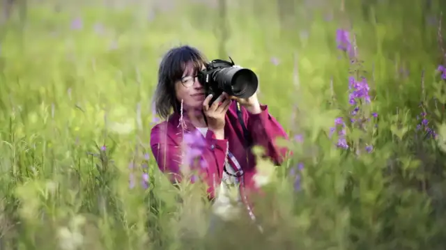 Kobieta w okularach pośród kwitnącej łąki robi zdjęcie aparatem z jasnym obiektywem, który pozwala uchwycić piękno natury.