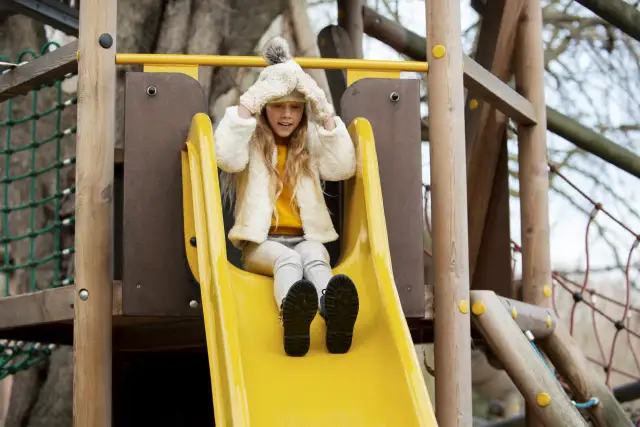 Dziewczynka w czapce z pomponem zjeżdża po żółtej zjeżdżalni na placu zabaw.