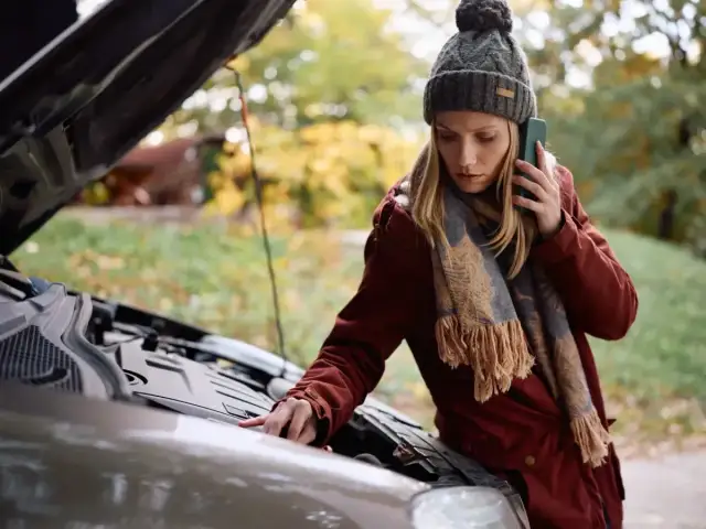 Kobieta z telefonem przy uchu sprawdza silnik auta, zastanawiając się, dlaczego nie można holować automatu.