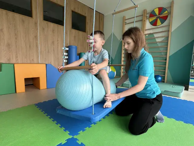 Terapeuta pomaga dziecku na huśtawce z piłką terapeutyczną w sali integracji sensorycznej.