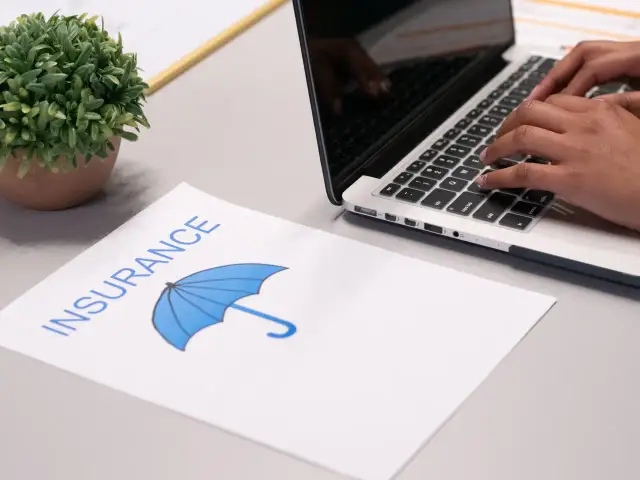 Osoba wpisuje dane na laptopie, obok leży dokument z napisem "INSURANCE" i symbolem parasola, sugerujący ubezpieczenie domu od wszystkich ryzyk.