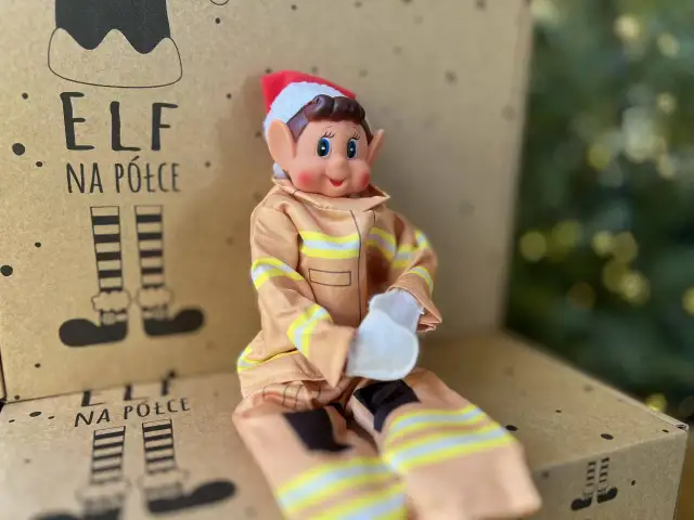 Uroczy elf na półce w stroju strażaka siedzi na kartonie z rysunkami elfich nóg.
