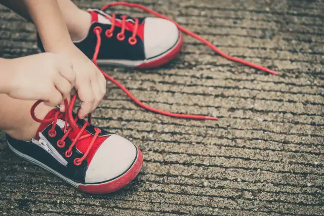 Dziecko wiążące sznurówki w czerwono-czarnych trampkach na betonowym podłożu.