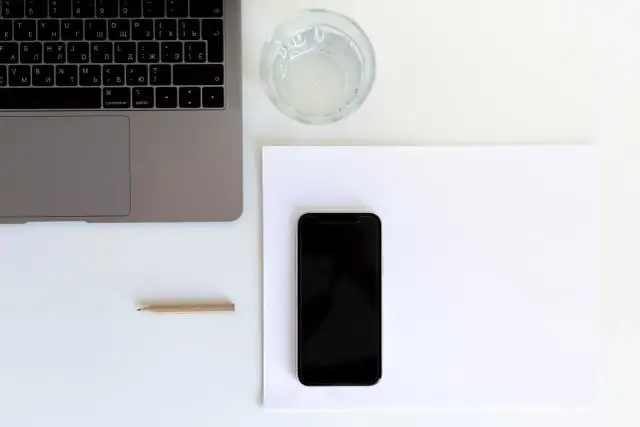 Przygotowanie do naklejania szkła na telefon. Obok laptopa i szklanki wody leży czarny smartfon na białej kartce.