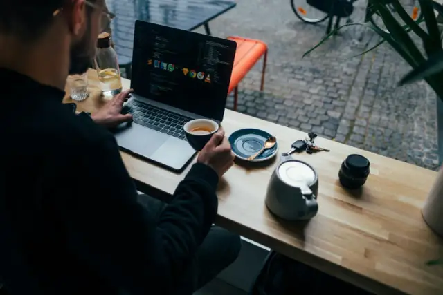 Mężczyzna pracuje na laptopie, popijając kawę. Widać kod na ekranie, klucze i aparat. To przykład multitasking co to znaczy być produktywnym.