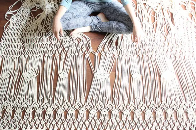 Osoba siedzi na podłodze, tworząc skomplikowane makrama wzory. Widać misternie splecione sznurki, tworzące geometryczne kształty.