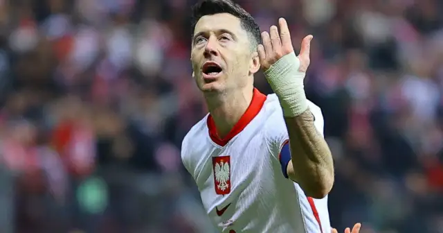 Robert Lewandowski w koszulce reprezentacji Polski, z bandażem na ręce, wznosi gest ku górze. Fani zastanawiają się, o której mecz piłki nożnej.