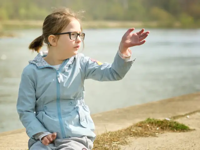 Dziewczynka z niepełnosprawnością w okularach siedzi nad wodą, wyciągając rękę.