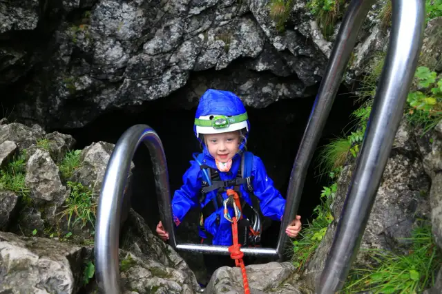 Dziecko w kasku i niebieskiej kurtce zjeżdża po poręczy w skalnym przejściu.