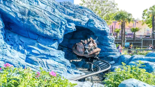 Personas en un coche de montaña rusa saliendo de una cueva azul rocosa, con palmeras y un paisaje de cuento de hadas al fondo.