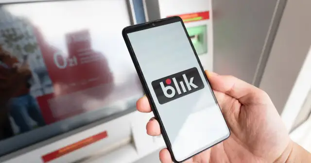 Dłoń trzyma smartfon z logo BLIK przed bankomatem.