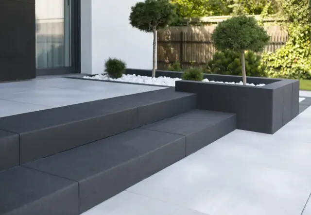 Nowoczesne schody z ciemnych płyt chodnikowych przed domem z białymi płytkami i zielenią.