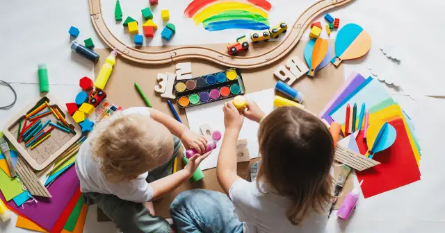 Dzieci tworzą kolorowe prace, jakby planowały, jak zdobyć dofinansowanie do przedszkola. Widać klocki, farby, kredki i papier.
