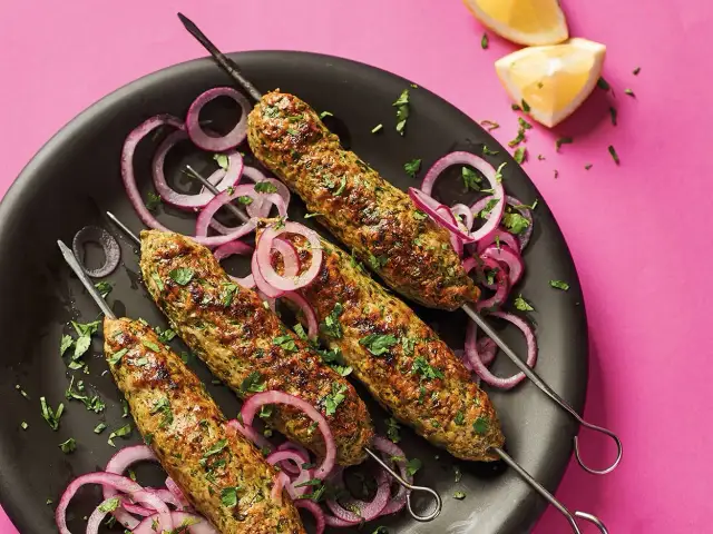 Cztery szaszłyki z mielonego mięsa z ziołami, podane na czarnym talerzu z krążkami czerwonej cebuli i posiekaną natką pietruszki.