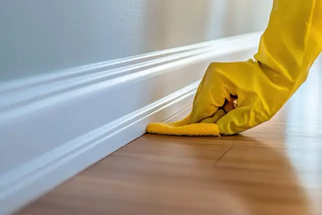 Dłoń w żółtej rękawiczce czyści białą listwę przypodłogową na drewnianej podłodze.