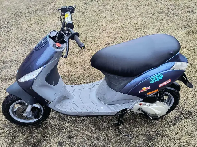 Niebieski skuter z naklejkami Red Bull i Motul na tle trawy.