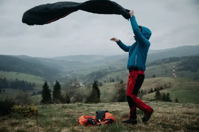 Osoba w niebieskiej kurtce i czerwonych spodniach rozkłada śpiwór puchowy czy syntetyczny na tle górskiego krajobrazu.