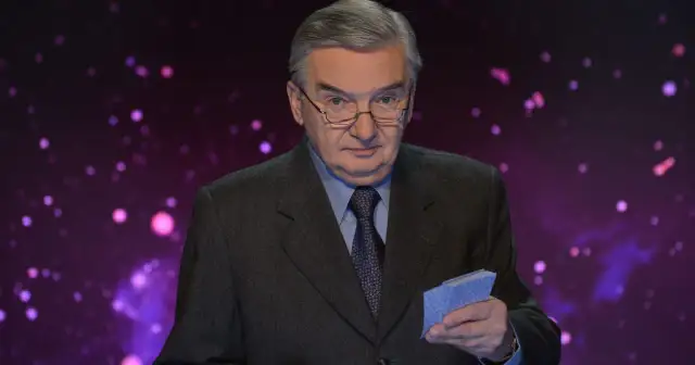 Tadeusz Sznuk w garniturze i okularach trzyma w ręku niebieskie karty na tle fioletowej, rozmytej scenografii.