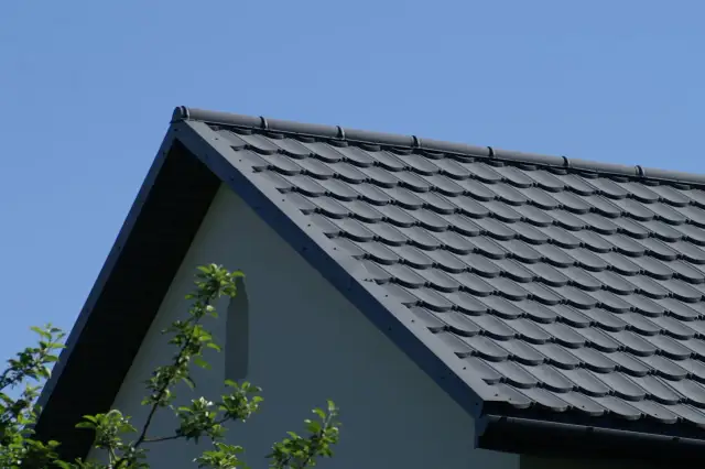 Szary dach z dachówki ceramicznej pod błękitnym niebem, z fragmentem ściany domu i gałęziami drzew.