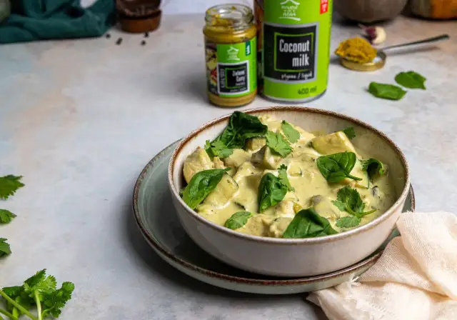 Miseczka zielonego curry z kurczakiem i szpinakiem, obok puszka mleka kokosowego i słoiczek pasty curry.