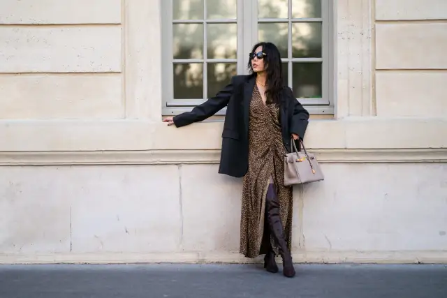 Elegancka kobieta w sukience w panterkę i czarnym blazerze, z beżową torebką i brązowymi kozakami dla niskiej osoby, które optycznie wydłużają nogi.