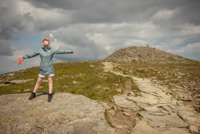 Kobieta z rozłożonymi rękami na szczycie Babiej Góry. Takie to góry, surowe i piękne, z kamienistym szlakiem i chmurami na niebie.