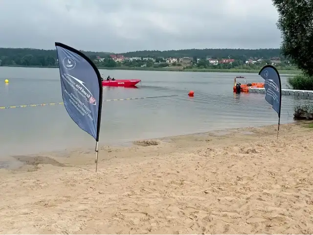 Plaża nad jeziorem, czerwona łódź ratownicza WOPR, pomarańczowa łódź, flagi, budynki na horyzoncie.