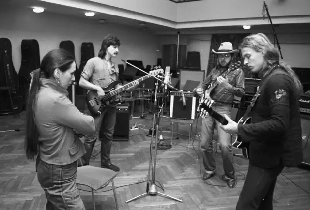 Zespół muzyczny w studiu, w tle stojaki na instrumenty. Wokalistka czeka na wiatr co rozgoni, a muzycy grają na gitarach.
