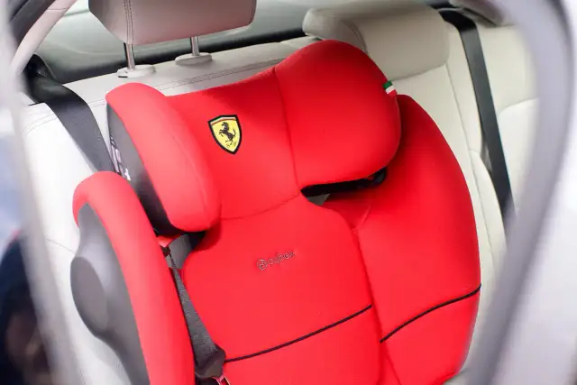Czerwony fotelik samochodowy Cybex z logo Ferrari. Pasek bezpieczeństwa przechodzi przez fotelik, pokazując, jak prawidłowo zapinać fotelik samochodowy.