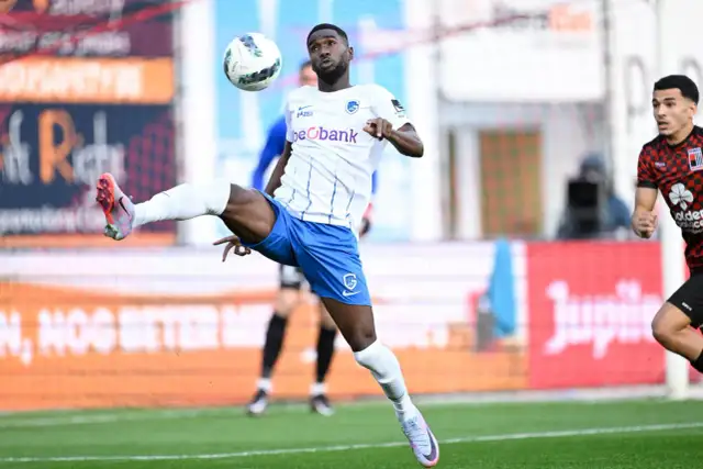 Piłkarz w białej koszulce i niebieskich spodenkach wykonuje akrobatyczne zagranie piłką na boisku.