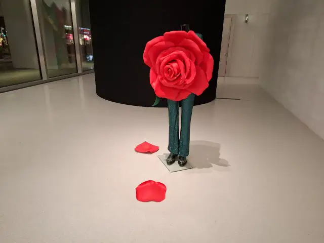 Manekin w błyszczących spodniach i wielka czerwona róża na tle czarnej ściany.