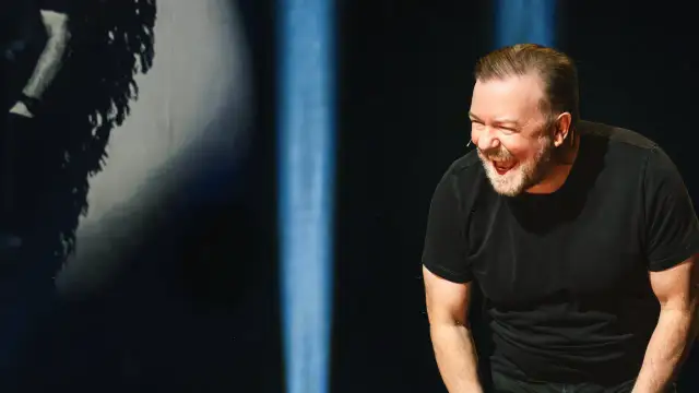 Ricky Gervais w swoim stand upie śmieje się do rozpuku, w czarnym t-shircie, na tle niebieskich świateł.