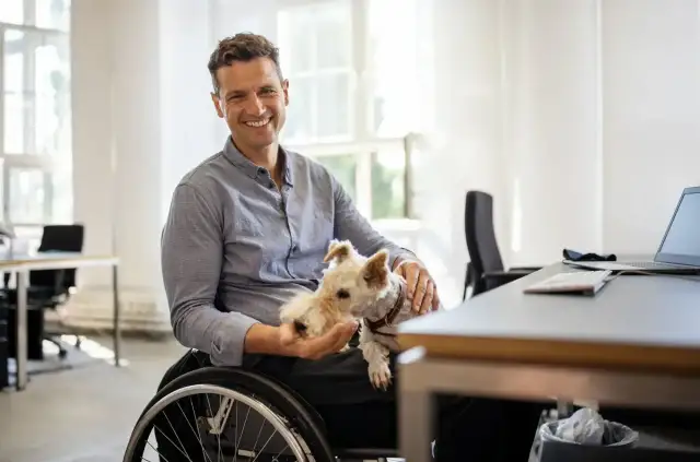 Uśmiechnięty mężczyzna na wózku inwalidzkim trzyma na rękach małego, białego psa w biurze.