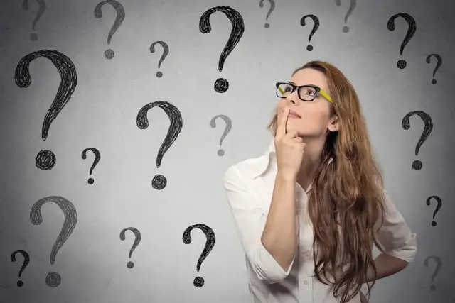 Kobieta w okularach z palcem przy ustach, otoczona znakami zapytania.