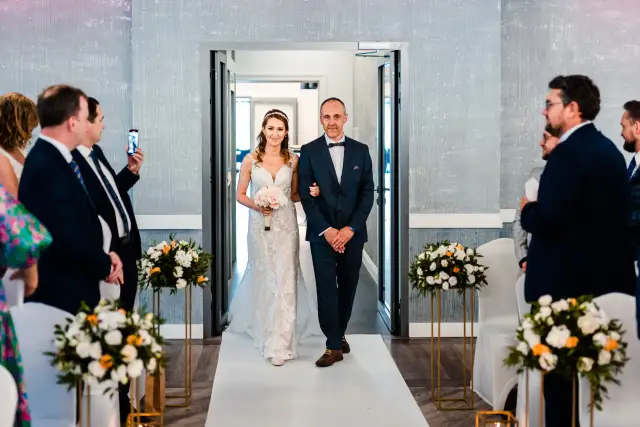 Panna młoda w sukni ślubnej idzie z ojcem do ołtarza, obok bukiety kwiatów.