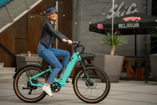 Uśmiechnięta kobieta w kasku i jeansach na turkusowym rowerze elektrycznym. Idealny wybór dla osób szukających dofinansowania do roweru elektrycznego.
