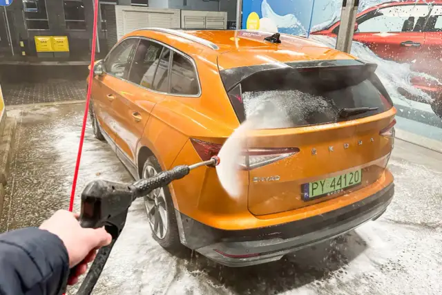 Pomarańczowy Skoda Enyaq jest myty na myjni bezdotykowej. Woda pod ciśnieniem spływa po karoserii, tworząc pianę.