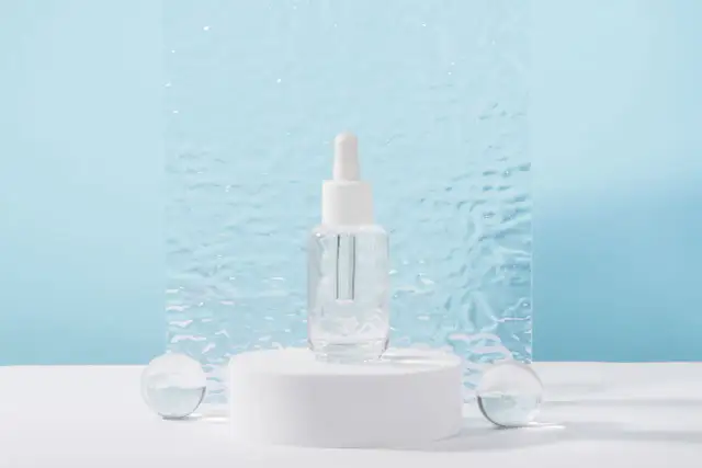 Szklana buteleczka z pipetką, zawierająca serum z witaminą B3, na białym postumencie, w otoczeniu szklanych kul.