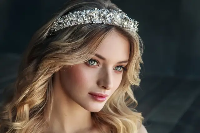 Piękna kobieta z blond włosami i hipnotyzującymi oczami, ozdobiona bogato zdobionym diademem, który przypomina tiarę.