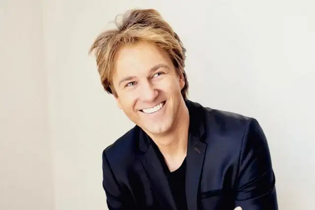 Uśmiechnięty mężczyzna w czarnej marynarce, przypominający Kaszewiaka, gotowy do stand-upu.