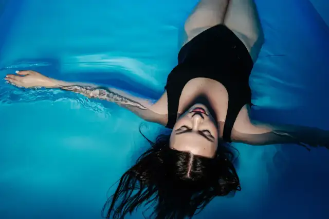 Kobieta w czarnym kostiumie kąpielowym floating, z zamkniętymi oczami, rozłożonymi ramionami, unosi się na wodzie.