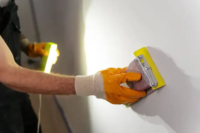 Ręka w pomarańczowej rękawicy szlifuje ścianę papierem ściernym. To idealny sposób, czym szlifować ściany, by uzyskać gładką powierzchnię.