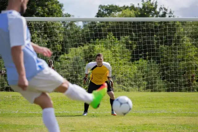 Piłkarz strzela w kierunku bramkarza stojącego przed bramką.